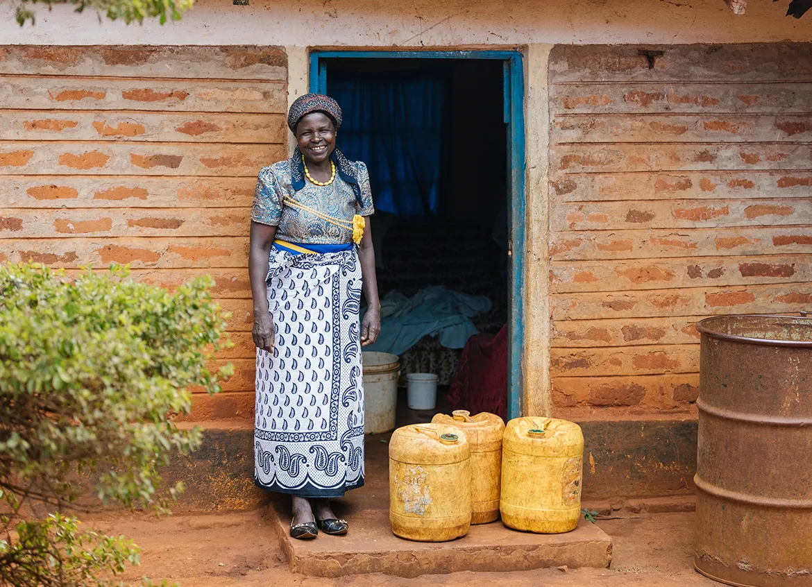 A woman smiling in front of her home next to 3 yellow water vessels.